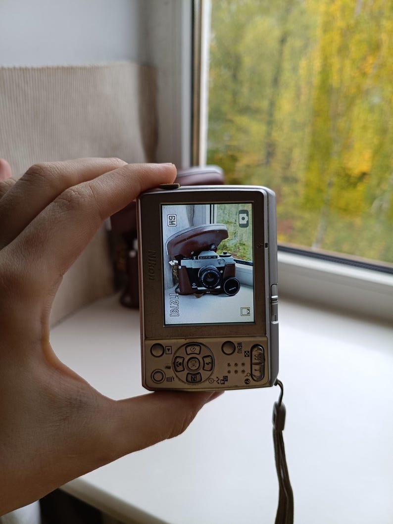 May include: A silver Nikon digital camera with a rectangular body, a rear LCD screen displaying a photograph of a vintage film camera in a brown leather case, a multi-directional navigation pad, and a wrist strap attached to the side.