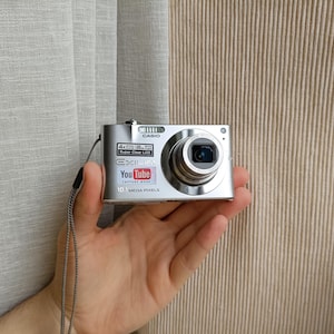 Może przedstawiać: Srebrny aparat cyfrowy Casio Exilim z obiektywem z 4-krotnym zoomem optycznym, matrycą 10,1 megapiksela oraz naklejką YouTube capture mode z przodu. Kompaktowe urządzenie posiada pasek na nadgarstek oraz przycisk migawki.