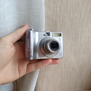 Puede incluir: Una cámara digital plateada Canon PowerShot A550 sostenida en una mano. La cámara tiene una lente, flash y varios botones. El texto "Canon" es visible en el lateral. La cámara mide aproximadamente 9.4 cm de largo.