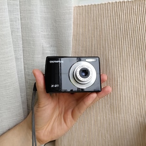 May include: A black Olympus X-21 digital camera featuring a silver lens barrel with Olympus Lens 3X Optical Zoom 5.8-17.4mm 1:2.8-4.8 text printed on the front. The compact camera includes a small flash unit and a grey wrist strap attached to the side.
