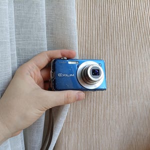 Może przedstawiać: Niebieski aparat cyfrowy Casio Exilim ze srebrnym obiektywem, metalowym srebrnym przyciskiem migawki i paskiem na nadgarstek. Na przedniej części obudowy aparatu znajduje się biały nadruk z nazwą marki Casio oraz modelem Exilim.