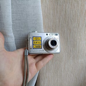 May include: A silver Sony Cyber-shot digital camera with a wrist strap. The front features a lens and a yellow sticker displaying technical specifications including 7.2 megapixels, a 2.4 inch LCD monitor, and ISO 1000 high sensitivity settings.