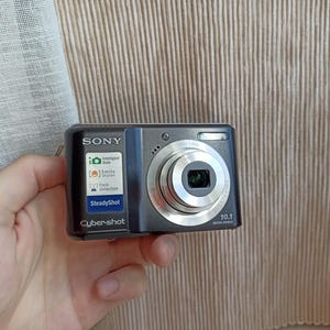 Puede incluir: Cámara digital Sony Cyber-shot negra con lente plateada y texto "10.1 MEGA PIXELS". Incluye funciones como Intelligent Auto, Smile Shutter, Face Detection y SteadyShot.