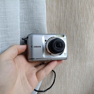 以下が含まれることがあります： 3.3倍光学ズームレンズ、内蔵フラッシュ、10.0メガピクセルセンサーを搭載したシルバーのキヤノン製デジタルカメラ。コンパクトな本体の側面には黒いリストストラップが付いており、前面にはCanonのブランド名が印字されています。