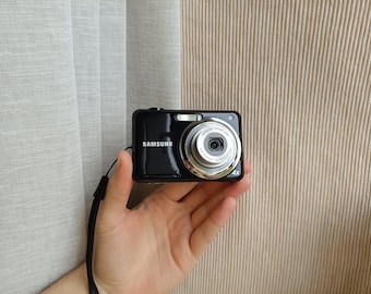 May include: A black Samsung digital camera featuring a silver lens barrel with 4x zoom capabilities, a built-in flash, and a black wrist strap. The lens housing displays technical text including Samsung Zoom Lens 4.9-19.6mm 1:2.9-6.5 27mm.