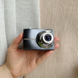 Puede incluir: Una cámara digital compacta de color gris oscuro con un barril de lente plateado que muestra el texto Kodak AF 3x Optical Aspheric Lens 36mm-108mm Equivalent. El cuerpo incluye un flash rectangular sobre el lente y una fina línea plateada.