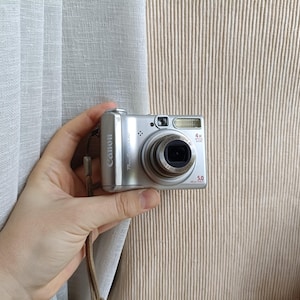 以下が含まれることがあります： 4倍光学ズームレンズ、内蔵フラッシュ、5.0メガピクセルセンサーを搭載したシルバーのキヤノンPowerShotデジタルカメラ。コンパクトカメラには側面にリストストラップが取り付けられており、前面にキヤノンのブランド名が表示されています。