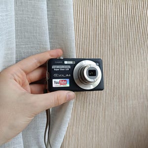 May include: A black Casio Exilim digital camera featuring a 10.1 megapixel Super Clear LCD and a YouTube Capture Mode sticker on the front. The camera has a silver lens barrel and includes a thin wrist strap attached to the side of the device.