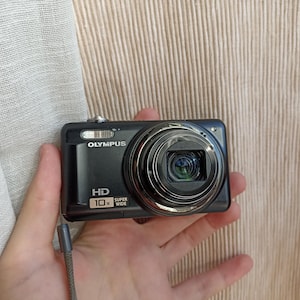 Peut inclure: Appareil photo numérique Olympus noir avec un objectif zoom 10x. Les mots "HD" et "SUPER WIDE" sont imprimés sur le devant. L'appareil photo est tenu dans une main.