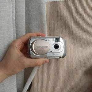 Puede incluir: Una cámara digital plateada Olympus Camedia D-395 con un sensor de 3,2 megapíxeles y una tapa de lente circular. La parte frontal del dispositivo muestra el nombre de la marca, el número de modelo y las especificaciones técnicas en texto negro y rojo.