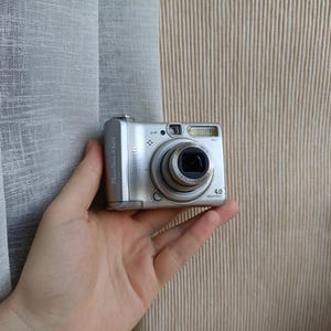 以下が含まれることがあります： 4.0メガピクセルのレンズ、内蔵フラッシュ、目立つズームレンズバレルを備えたシルバーのキヤノンPowerShot A520デジタルカメラ。カメラ本体の側面にはモデル名のPowerShot A520、前面には4.0 Megapixelsの文字が表示されています。