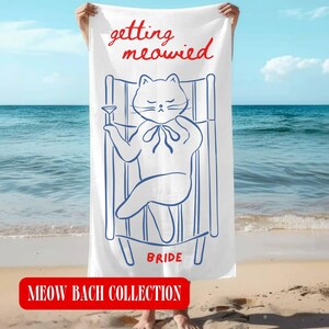 May include: A white beach towel featuring a blue line drawing of a cat lounging in a deck chair while holding a cocktail glass. The text "getting meowied" appears in red above the cat, and the word "BRIDE" is printed in red below the chair.