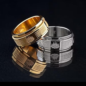 K&ouml;nnte beinhalten: Zwei Ringe, einer goldfarben und einer silberfarben, werden auf einer reflektierenden Oberfl&auml;che pr&auml;sentiert. Beide Ringe weisen ein eingraviertes Design mit einem zentralen Emblem und linearen Mustern auf. Der goldfarbene Ring ist im Vordergrund.