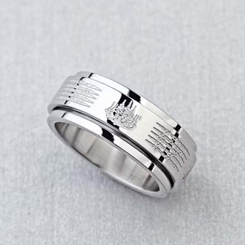 K&ouml;nnte beinhalten: Ein silberfarbener Ring mit ge&auml;tzten Mustern und einem drehbaren Au&szlig;enband. Der Ring weist detaillierte Designs auf, m&ouml;glicherweise religi&ouml;ser oder kultureller Natur, was eine einzigartige &Auml;sthetik verleiht. Der Ring wird auf einer hellgrauen Oberfl&auml;che gezeigt.