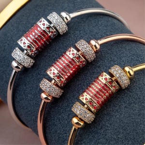 May include: Three bangle bracelets with decorative red and gold centerpieces. The centerpieces feature a red enamel design with heart and cross details, accented by sparkling clear stones. The bracelets are in silver, rose gold, and gold tones.