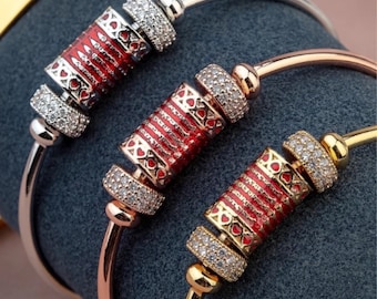 May include: Three bangle bracelets with decorative red and gold centerpieces. The centerpieces feature a red enamel design with heart and cross details, accented by sparkling clear stones. The bracelets are in silver, rose gold, and gold tones.