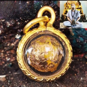 May include: A gold-colored pendant with a circular design. The pendant features a clear, rounded center filled with gold leaf. The outer frame has a decorative, textured pattern. A small loop is at the top for a chain. The background is dark and blurred.