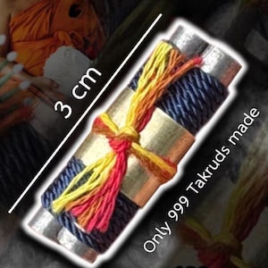 May include: A cylindrical metal amulet wrapped in dark blue cord and tied with yellow, orange, and red thread. The object is marked with 3 cm and the text Only 999 Takruds made. The metallic surface is visible between the decorative cord wrappings.