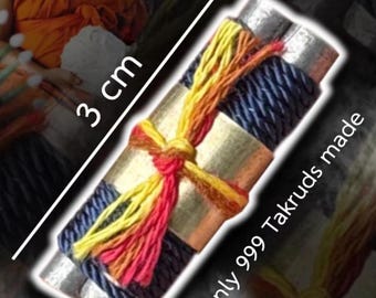 May include: A cylindrical metal amulet wrapped in dark blue cord and tied with yellow, orange, and red thread. The object is marked with 3 cm and the text Only 999 Takruds made. The metallic surface is visible between the decorative cord wrappings.