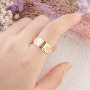 K&ouml;nnte beinhalten: Ein goldfarbener Ring mit strukturiertem Band und kleinem Emblem. Der Ring wird an einem Finger getragen, wodurch sein Design hervorgehoben wird. Das Design des Rings ist schlicht, aber elegant und f&uuml;r den Alltag geeignet. Der Ring ist wahrscheinlich aus Gold.