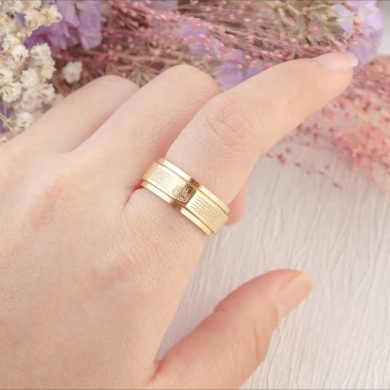 K&ouml;nnte beinhalten: Ein goldfarbener Ring mit strukturiertem Band und kleinem Emblem. Der Ring wird an einem Finger getragen, wodurch sein Design hervorgehoben wird. Das Design des Rings ist schlicht, aber elegant und f&uuml;r den Alltag geeignet. Der Ring ist wahrscheinlich aus Gold.