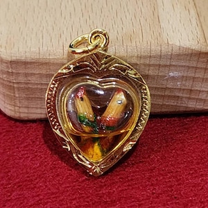 May include: A gold-coloured pendant with a heart-shaped frame. Inside, two small, painted wooden objects are visible, encased in clear material. The pendant has a loop for a chain and rests on a red surface.