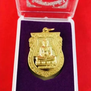 Puede incluir: Colgante dorado con una figura de Buda sentado dentro de un marco en forma de escudo. El colgante se exhibe en una caja de plástico transparente con un respaldo de terciopelo púrpura, sobre un fondo rojo.