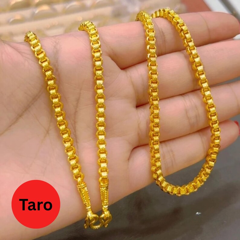 May include: A gold-colored chain necklace with a box link design. The necklace is held in a hand, and the word "Taro" is in a red circle in the lower left corner.