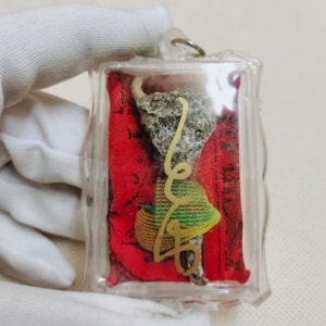 Op de afbeelding: Een heldere, rechthoekige hanger met een klein, getextureerd grijs object, een geweven mand en een groen en gele hoed, tegen een rode achtergrond. Een goudkleurig, gestileerd script is ook zichtbaar.