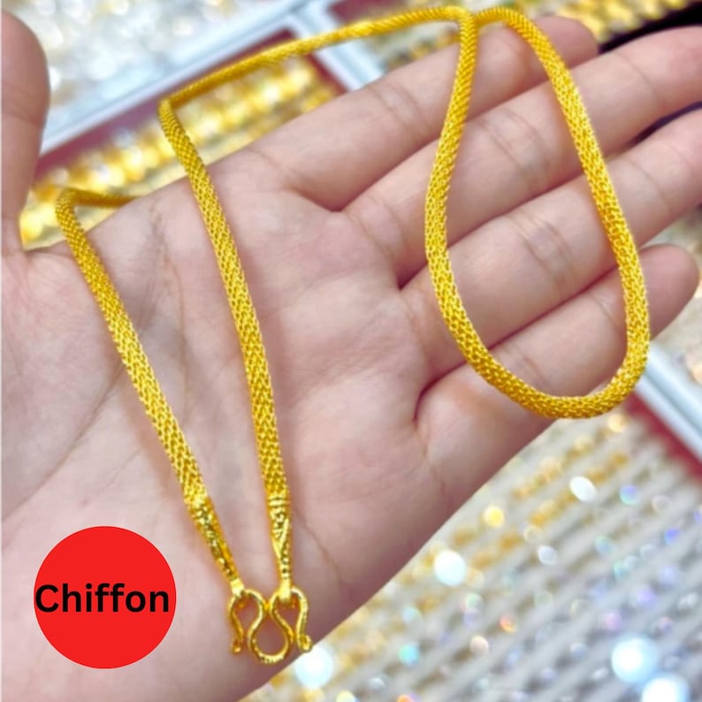 May include: A gold-colored necklace with a woven chain design. The necklace is held in a hand, and the clasp is visible. The word "Chiffon" is in a red circle in the lower left corner.