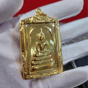May include: A gold pendant with a seated Buddha figure inside a rectangular frame. The pendant has a detailed border and a loop for a chain. The Buddha is shown in a meditative pose, with intricate detailing.