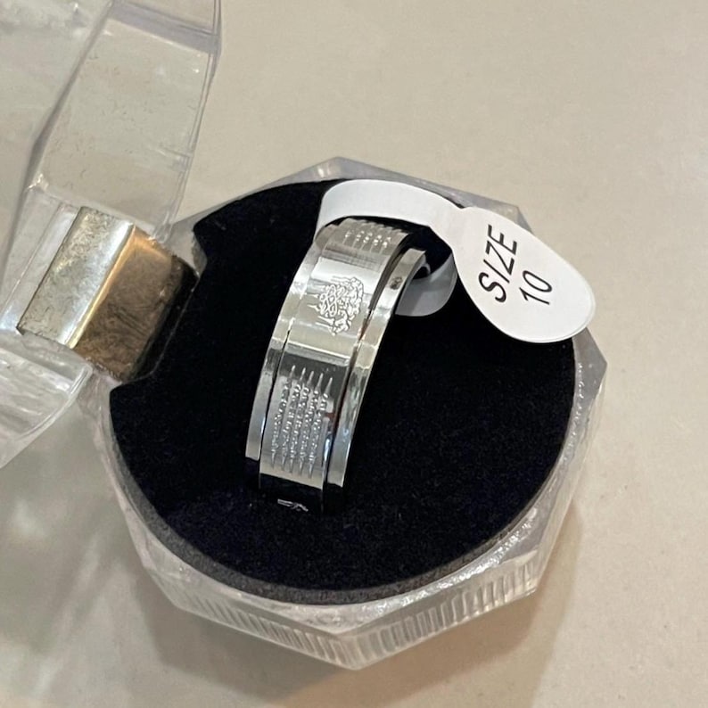 K&ouml;nnte beinhalten: Ein silberner Ring mit aufwendigen Details, pr&auml;sentiert in einer transparenten Kunststoffbox mit schwarzem Samtinnenraum. Der Ring hat ein Gr&ouml;&szlig;enetikett 10. Das Design des Rings umfasst eingravierte Muster und m&ouml;glicherweise Schrift.