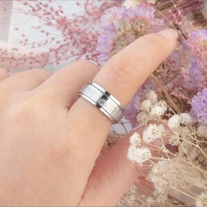 K&ouml;nnte beinhalten: Ein silberfarbener Ring mit strukturiertem Band und einem kleinen, dunklen Emblem. Der Ring wird an einem Finger getragen, mit einem verschwommenen floralen Hintergrund. Das Design des Rings umfasst eine Reihe paralleler Linien und ein zentrales dekoratives Element.
