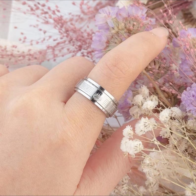 K&ouml;nnte beinhalten: Ein silberfarbener Ring mit strukturiertem Band und einem kleinen, dunklen Emblem. Der Ring wird an einem Finger getragen, mit einem verschwommenen floralen Hintergrund. Das Design des Rings umfasst eine Reihe paralleler Linien und ein zentrales dekoratives Element.