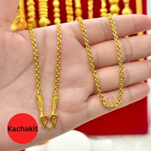 May include: A gold-colored necklace with a delicate chain design. The necklace is held in a hand, with a red circle that says "Kachakit" in white text. The necklace has a decorative clasp.