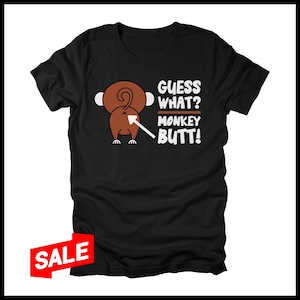 May include: Black t-shirt featuring a cartoon monkey's rear end with the text "Guess What? Monkey Butt!" in white. The monkey is brown with a white arrow pointing to its rear. A red "SALE" tag is in the lower left corner.