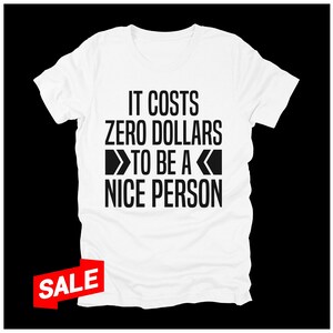 May include: White t-shirt with the text "IT COSTS ZERO DOLLARS TO BE A NICE PERSON" in black. A red and white "SALE" tag is in the lower left corner. The shirt is displayed against a black background.