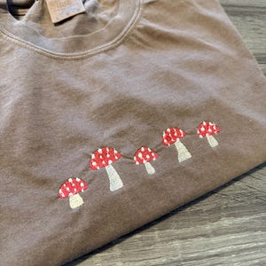 May include: Brown t-shirt with a crew neck and embroidered red and white mushroom design. The mushrooms are arranged in a row across the front of the shirt. The shirt is folded.