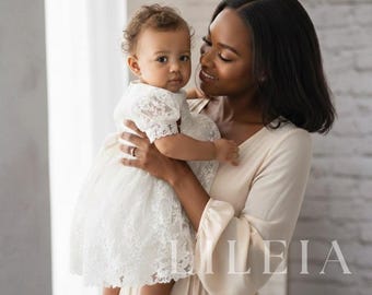 May include: A white infant dress featuring intricate floral lace overlays, short puff sleeves, and a scalloped hemline. The dress is layered over a solid white fabric. The text LILEIA is visible in the lower right corner of the image.