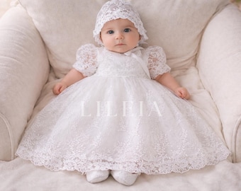May include: A baby wearing a white lace christening gown and bonnet, seated on a white upholstered chair. The dress features short, puffy sleeves and a delicate floral lace pattern. The word "LILIEIA" is printed on the dress.