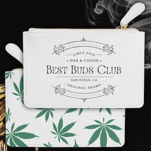 Könnte beinhalten: Weiße Reißverschlusstasche mit goldfarbenem Reißverschluss und Zuglasche. Auf der Tasche steht in Schwarz "BEST BUDS CLUB" und "SINCE 2025". Die untere Tasche hat ein grünes Cannabisblattmuster.