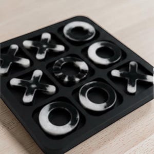 May include: A black tic-tac-toe game board featuring a grid of nine square slots. The game pieces consist of black and white marbled X and O shapes placed within the slots, creating a high-contrast visual pattern across the square game board.