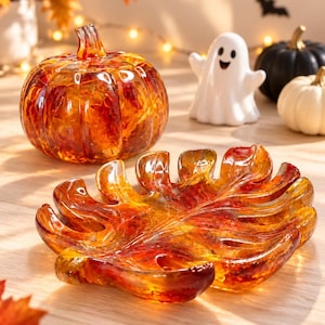 May include: A glass pumpkin figurine and a matching leaf-shaped glass dish, both featuring a translucent amber, orange, and red marbled pattern. The pumpkin has a rounded, ribbed body with a stem, while the dish mimics a large tropical leaf.