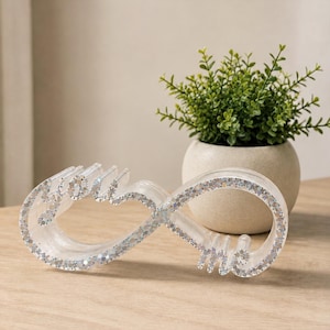 May include: A clear, infinity-shaped decorative object made of resin with embedded silver glitter. The cursive text "you" is integrated into the left loop, and the word "me" is integrated into the right loop of the infinity symbol.