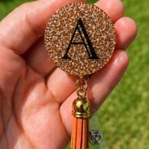 May include: A circular badge reel featuring a gold glitter finish and a prominent black letter A in a serif font. The badge reel is attached to a gold-toned metal connector and a short, orange-coloured tassel that hangs vertically from the base.