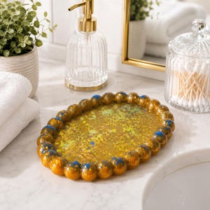 May include: An oval-shaped tray with a beaded border, featuring a gold and blue speckled finish with iridescent flakes embedded in the center. The tray has a glossy, reflective surface and a textured, rounded edge design.