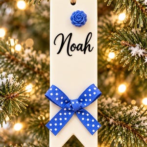 Puede incluir: Un adorno rectangular de cerámica blanca con un borde inferior en forma de muesca, que presenta el nombre Noah en letra cursiva negra, un pequeño adorno floral azul en la parte superior y un lazo azul con lunares blancos atado en la base.