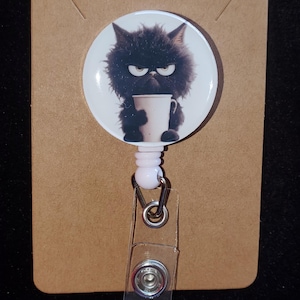 May include: A retractable badge reel featuring a grumpy black cat holding a white mug. The cat has a stern expression. The reel is attached to a clear plastic clip and a pink reel. The background is a brown card.