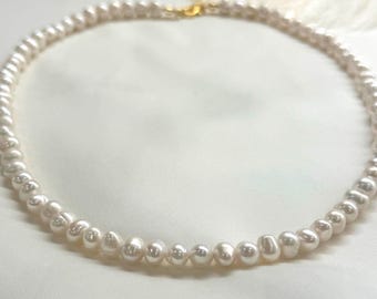 May include: A delicate pearl necklace with a gold clasp. The necklace features a strand of lustrous, off-white pearls, varying slightly in size. The pearls are strung closely together, creating a smooth, elegant look. The background is a soft, white fabric.