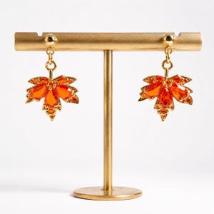 May include: A pair of gold-toned drop earrings featuring a maple leaf design. Each earring is adorned with multiple faceted orange crystals arranged in a leaf shape, suspended from a small gold-toned ball stud. The earrings are displayed on a gold stand.